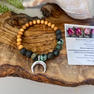 May include: A beaded bracelet featuring wooden beads, black lava beads, and green-speckled stone beads. A silver crescent moon charm hangs from the center. The bracelet rests on a wooden surface, with a business card in the background.