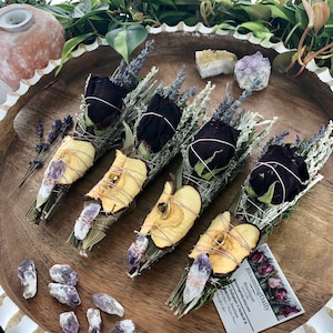 Op de afbeelding: Vier bundels gedroogde kruiden en bloemen gewikkeld in gedroogde appelschijfjes en vastgebonden met touw. Elke bundel bevat gedroogde lavendel, salie en een zwarte roos. Amethistkristallen zijn rond de bundels verspreid. De tekst op de kaart luidt "Blooming Daily Boutique".