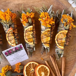 May include: Several sage bundles with dried orange slices, cinnamon sticks, and bright orange flowers are arranged on a wooden tray. The bundles are wrapped with black string. A small card with text is also present.