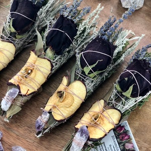 Op de afbeelding: Vier wierookstokjes met gedroogde appelschijfjes, zwarte rozen, lavendel en salie. Elk wierookstokje is vastgebonden met touw en bevat een klein amethistkristal. De wierookstokjes zijn gerangschikt op een houten oppervlak met een klein visitekaartje waarop "Blooming Daily Boutique" staat.
