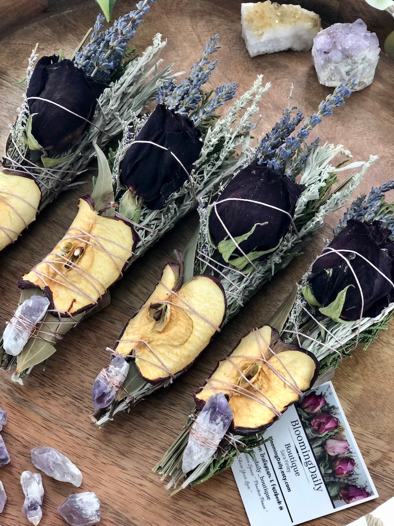 Op de afbeelding: Vier wierookstokjes met gedroogde appelschijfjes, zwarte rozen, lavendel en salie. Elk wierookstokje is vastgebonden met touw en bevat een klein amethistkristal. De wierookstokjes zijn gerangschikt op een houten oppervlak met een klein visitekaartje waarop "Blooming Daily Boutique" staat.