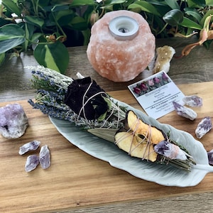 Op de afbeelding: Een wit keramiek bord in de vorm van een veer bevat een wierookstokje met gedroogde lavendel, salie, rozenblaadjes en appelschijfjes. Het wierookstokje is vastgebonden met touw en ligt op een houten oppervlak met amethistkristallen en een Himalaya-zoutlamp.