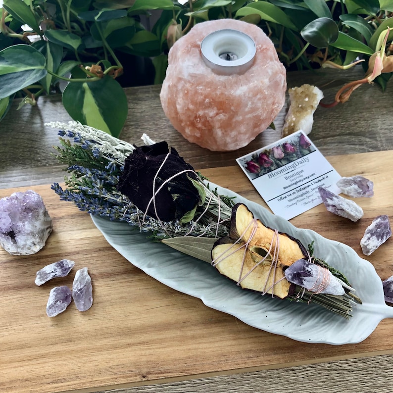 Op de afbeelding: Een wit keramiek bord in de vorm van een veer bevat een wierookstokje met gedroogde lavendel, salie, rozenblaadjes en appelschijfjes. Het wierookstokje is vastgebonden met touw en ligt op een houten oppervlak met amethistkristallen en een Himalaya-zoutlamp.