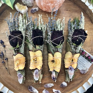 Op de afbeelding: Vier bundels gedroogde kruiden en bloemen gebonden met touw, waaronder lavendel, salie en gedroogde appelschijfjes. Elk bundel is bedekt met een zwarte roos en een amethistkristal. De bundels zijn gerangschikt op een houten oppervlak.