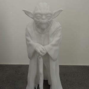 Peut inclure: Figurine blanche de Yoda, personnage de la franchise Star Wars, debout avec une canne. La figurine porte une robe et a de grandes oreilles. La figurine est sur une surface gris foncé.