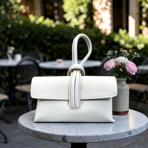 May include: A white leather clutch with a knotted handle. The rectangular bag has a flap closure and a textured surface. The handle is looped and knotted at the top. A small vase with pink and white flowers sits nearby on a marble table.