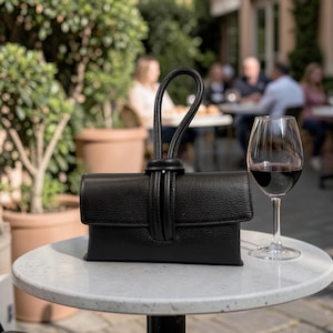 May include: A black leather clutch bag with a looped handle rests on a white table beside a glass of red wine. The bag features a flap closure and a textured finish. The background shows a blurred outdoor cafe setting.