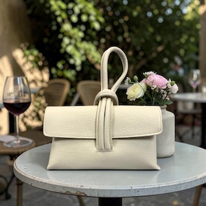 May include: Cream-colored leather handbag with a top handle and a flap closure. The bag has a textured surface and a knotted handle detail. A small vase of flowers sits nearby on a round table. A glass of red wine is in the background.