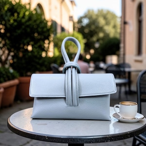 May include: A light blue leather clutch purse with a looped handle. The purse has a rectangular shape with a flap closure. A small white cup of coffee sits on a saucer next to the purse on a round table.