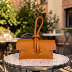 Könnte beinhalten: Eine hellbraune Leder-Clutch mit Schlaufengriff. Die rechteckige Tasche hat einen Klappenverschluss und eine strukturierte Oberfläche. Der Griff ist mit einem vertikalen Riemen an der Vorderseite befestigt. Die Tasche liegt auf einem runden Tisch.