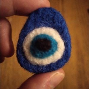 Peut inclure: Un porte-bonheur en feutre bleu en forme d'œil protecteur, avec un motif d'œil blanc, turquoise et noir. L'amulette de forme ovale est tenue entre les doigts. Le feutre est texturé.