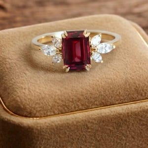 May include: A gold ring featuring a rectangular, deep red gemstone, with smaller white gemstones on either side. The ring is presented on a brown velvet box, with other decorative items on a wooden surface.