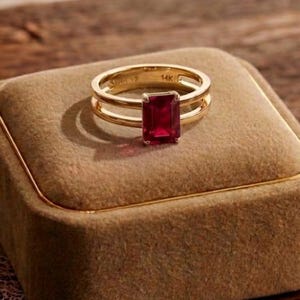 Peut inclure: Une bague en or avec une pierre précieuse rectangulaire rouge foncé est présentée sur un écrin en velours marron. La bague a un design à double bande. La boîte repose sur une surface en bois, avec d'autres objets décoratifs en arrière-plan.