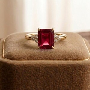 May include: A gold ring with a large, rectangular, red gemstone as the centerpiece. The ring has smaller, clear gemstones on either side of the main stone. The ring is displayed on a brown velvet ring box.