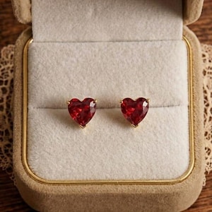 Puede incluir: Un par de pendientes de botón con piedras preciosas rojas en forma de corazón en una caja de joyería beige. Los pendientes están engarzados en metal dorado. La caja está abierta, revelando los pendientes contra un interior claro.