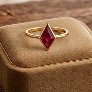 May include: A gold ring with a diamond-shaped, deep red gemstone. The ring is displayed on a square, tan velvet jewelry box with gold trim. The background is a wooden surface.