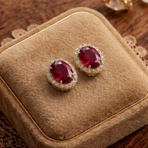 Può includere: Un paio di orecchini a lobo dorati con gemme ovali rosso scuro, circondate da un alone di pietre più piccole e trasparenti. Gli orecchini poggiano su un portagioie in velluto marrone con finiture dorate, su una superficie di legno.