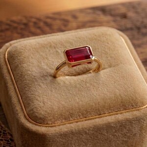 May include: A gold ring with a rectangular, deep red gemstone set in a gold band. The ring rests on a beige velvet ring box with gold trim. The background is a wooden surface with decorative elements.