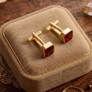 May include: A pair of gold-toned cufflinks with square, ruby-red gemstones, presented in a tan velvet jewelry box. The cufflinks have a classic design, perfect for formal wear. The box is rectangular with a gold trim.