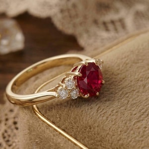 Könnte beinhalten: Ein Goldring mit einem runden, tiefroten Edelstein als Mittelstein, flankiert von drei kleinen, klaren, rund geschliffenen Akzentdiamanten auf jeder Seite. Die Ringschiene ist aus poliertem Gelbgold und hat eine klassische Krappenfassung.