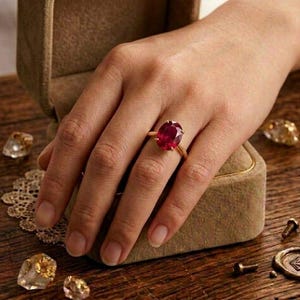 May include: A gold ring with a large, oval-cut ruby gemstone. The ring is on a person's finger, next to a velvet ring box. Other gemstones and decorative elements are scattered on a wooden surface.