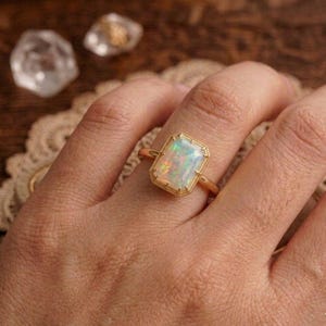 May include: A gold ring featuring a rectangular opal gemstone. The opal displays a play of color with flashes of green, blue, and orange. The ring has a delicate, beaded border around the gemstone.