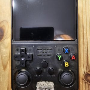 May include: A black handheld gaming console with a screen and control buttons. The console has a directional pad, two analog sticks, and action buttons labeled X, Y, A, and B. The text "OUAG Once Upon a Gamer" is visible on the console.