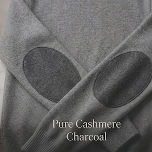 Puede incluir: Un suéter gris de cuello redondo con coderas color carbón. El suéter tiene una etiqueta que dice "Pure Cashmere". El texto "Pure Cashmere Charcoal" se muestra en la parte inferior de la imagen. El suéter está hecho de un material suave.