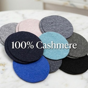 May include: A collection of round, 100% cashmere coasters in various colors, including blue, black, gray, and pink. The coasters are arranged on a marble surface, showcasing their soft texture and color variations.