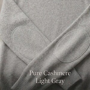 Può includere: Maglione in cashmere grigio chiaro con toppe sui gomiti. Il maglione ha un girocollo e polsini a coste. L'etichetta recita "Pure Cashmere". Il testo "Pure Cashmere Light Gray" è visualizzato nella parte inferiore dell'immagine.