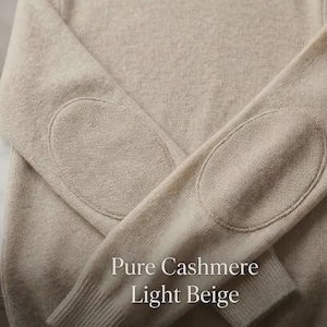 Peut inclure: Un pull en cachemire beige clair avec des coudières. Le pull a un col rond et des poignets côtelés. Le texte "Pure Cashmere Light Beige" est affiché en bas de l'image.