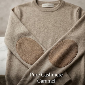 Puede incluir: Un suéter beige de cuello redondo con coderas marrones. El suéter tiene una etiqueta que dice "Pure Cashmere". El texto "Pure Cashmere Caramel" se muestra en la parte inferior de la imagen. El suéter está hecho de un material suave y cálido.