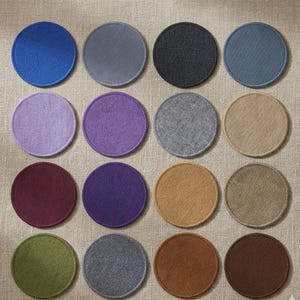 May include: A collection of sixteen round felt coasters in various colors, including blue, gray, purple, and brown. Each coaster has a stitched edge and is arranged on a neutral-colored fabric surface. These coasters are designed to protect surfaces from drink rings.