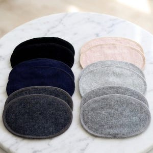 Peut inclure: Plusieurs coussinets d'allaitement ovales de différentes couleurs : noir, bleu marine, gris foncé, gris clair et rose. Les coussinets sont en matière douce et texturée, disposés en piles.