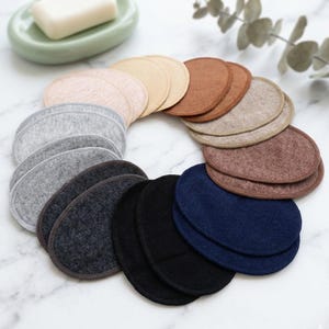 Peut inclure: Une collection de coussinets d'allaitement réutilisables de différentes couleurs, dont beige, gris, marron, noir et bleu marine. Les coussinets sont disposés en cercle, avec un pain de savon et un porte-savon en arrière-plan.