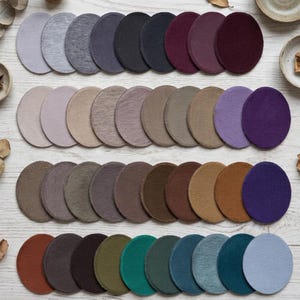 May include: A collection of round coasters in various colors, including shades of gray, purple, brown, and blue. The coasters are arranged in rows on a light-colored wooden surface. The coasters appear to be made of a soft, absorbent material.