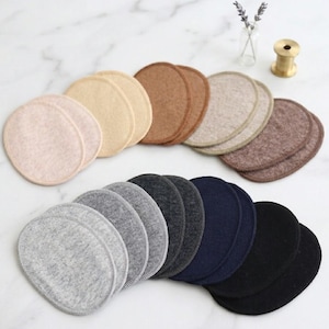 Op de afbeelding: Een verzameling ronde borstcompressen van stof in verschillende kleuren, waaronder beige, bruin, grijs, marineblauw en zwart. De pads zijn in paren gerangschikt en tonen hun vorm en textuur. Een kleine vaas met lavendel en een messing object staan op de achtergrond.