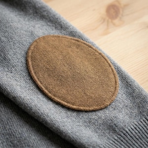 Op de afbeelding: Close-up van een grijze mouw van een trui met een bruine, ovale ellebooglap. De lap is op de mouw gestikt en biedt een contrast in textuur en kleur. De achtergrond is een lichtgekleurd houten oppervlak.