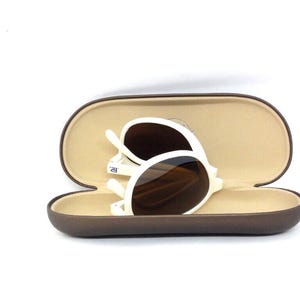 May include: White-framed sunglasses with brown lenses rest inside an open, brown oval case with a tan interior. The sunglasses are folded, showing the frame's design. The case is open, suggesting easy access.