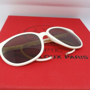 May include: White-framed sunglasses with dark lenses rest on a red box. The sunglasses have a classic, rounded shape. The box has the text "POUX PARIS" printed on it.
