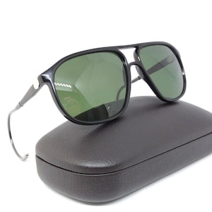 May include: Black aviator sunglasses with green lenses rest on a dark brown, textured case. The sunglasses have a double bridge and a sleek, modern design. The case is rectangular and appears to be made of leather or a similar material.