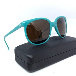May include: Teal-colored sunglasses with brown lenses rest on a black case. The sunglasses have a thick frame and a small logo on the temple. The lenses are a dark brown color, and the case is rectangular.