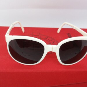 May include: White sunglasses with dark lenses are displayed on a red box. The sunglasses have a classic design with a thick white frame and curved arms. The lenses are dark, providing contrast against the white frame.