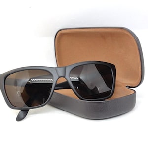 May include: Black sunglasses with dark lenses sit in an open, brown leather case. The sunglasses have a matte finish and a rectangular shape. The case is dark brown with a soft, suede-like interior.