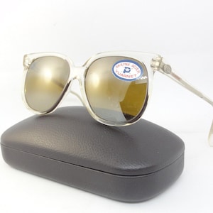 May include: Clear-framed sunglasses with gold-tinted lenses rest on a black case. The sunglasses have a square shape and a sticker on the lens. The case is rectangular with a textured surface. The sunglasses are a vintage style.