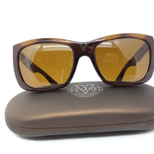 May include: Brown tortoiseshell sunglasses with amber lenses rest on a matching brown case. The sunglasses have a rectangular shape with a dark brown bridge and arms. The case has the brand name "Vuarnet Sunglasses" printed on it.