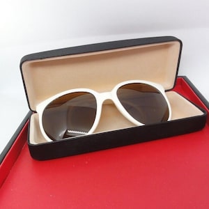 May include: White sunglasses with brown lenses rest in an open black case with a tan interior. The case sits on a red surface, creating a striking contrast. The sunglasses have a classic, rounded frame.