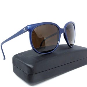 May include: Blue sunglasses with brown lenses rest on a black case. The sunglasses have a thick frame and a logo on the temple. The case is rectangular and has a matte finish. The sunglasses are a classic style.