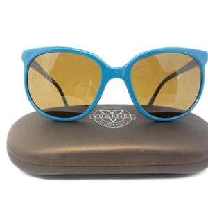 May include: Blue sunglasses with brown lenses rest on a brown case. The sunglasses have a thick, rounded frame. The case has a smooth, oval shape. The brand name "Vuarnet Sunglasses" is printed on the case.