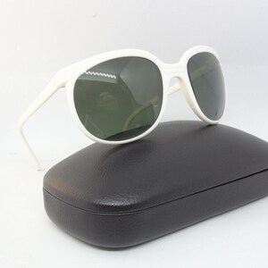 May include: White sunglasses with dark lenses rest on a black case. The sunglasses have a thick, rounded frame and a classic design. The case is rectangular with a slightly curved top. The overall aesthetic is vintage and stylish.